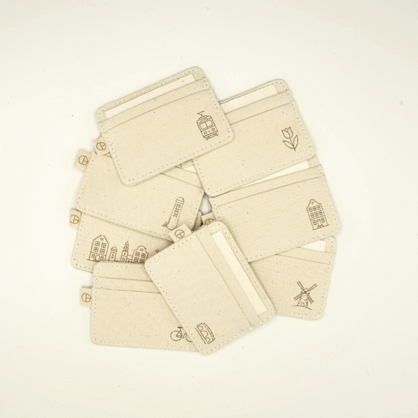 Overhead flatlay of 8 handmade CARDHEIR cardholders with engraved Amsterdam icons — including tram, tulip, windmill, bridge, and canal houses — crafted from sustainable natural canvas.