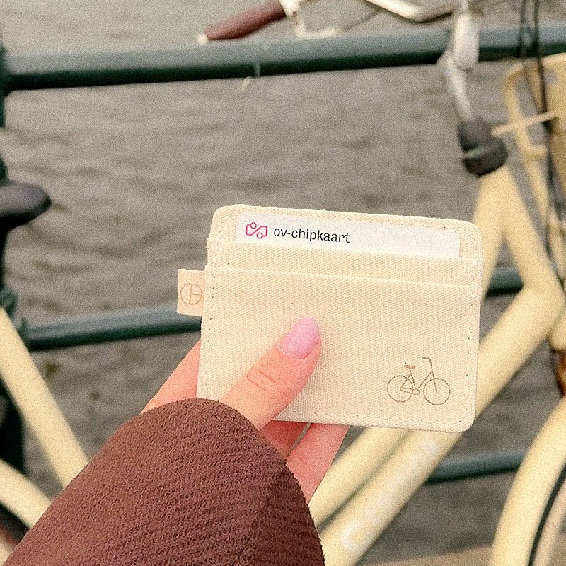 Hand holding a natural canvas CARDHEIR cardholder with a Omafiets icon, near an Amsterdam canal with a cream-colored bike in the background.