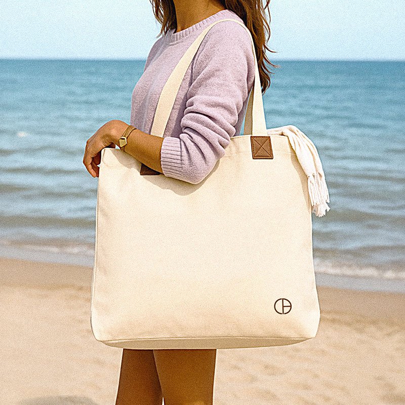 Woman holding a natural canvas CARDHEIR tote bag with the Kalani monogram, standing by the beach.