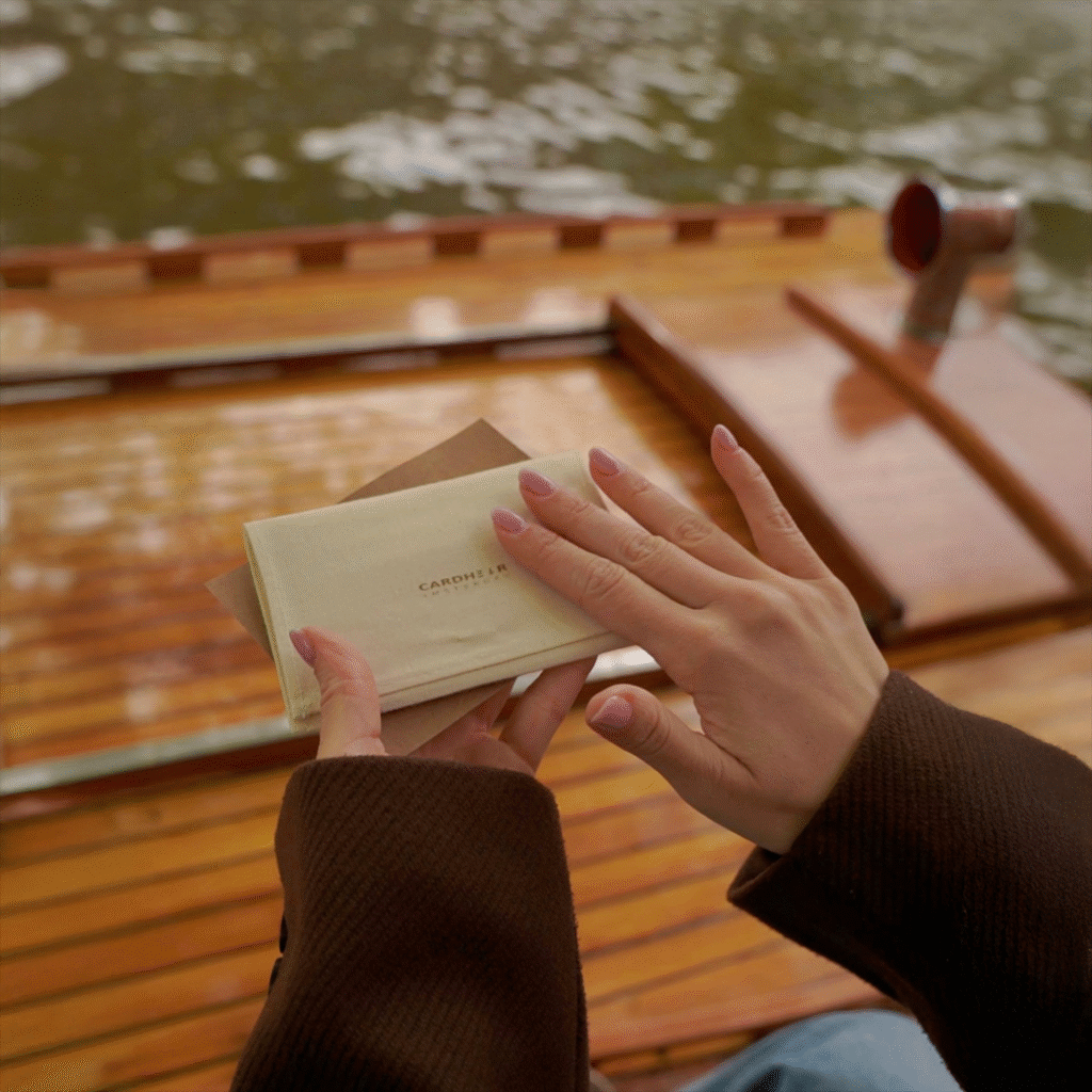 Hands gently holding a cotton pouch with the CARDHΞ↓R logo and a kraft envelope, above a wooden boat floor and canal water.