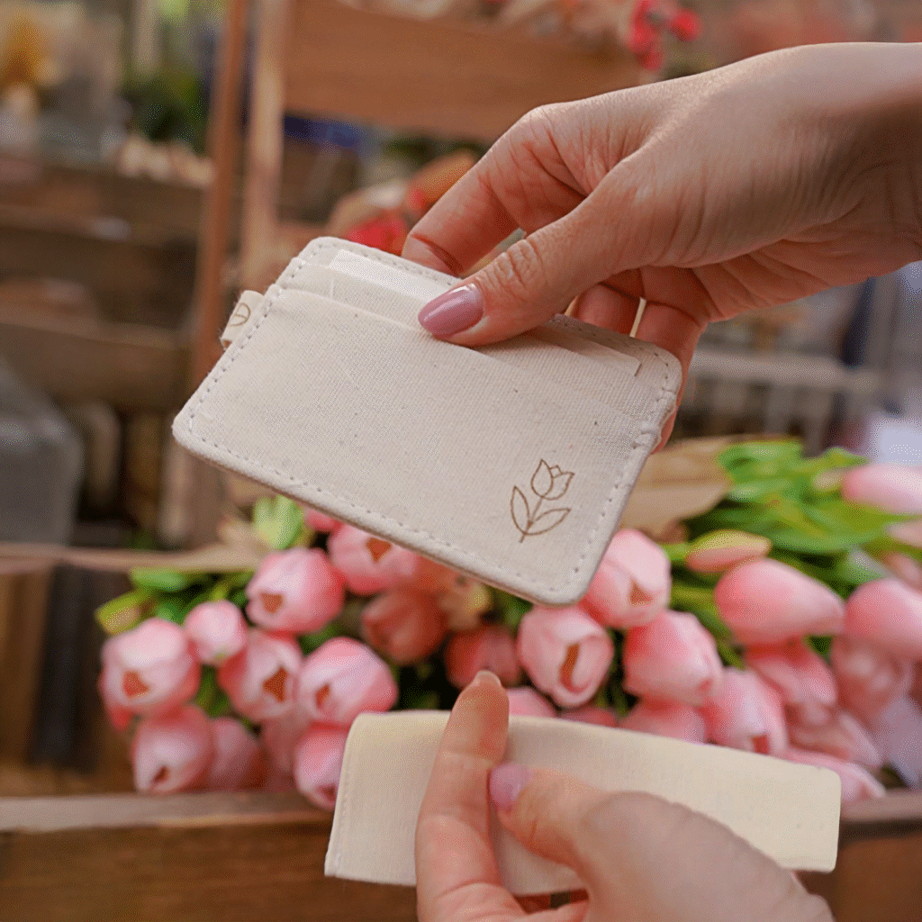 Hands opening a cotton pouch and holding a stitched cardholder with a tulip icon, with bunches of pink tulips in the background.