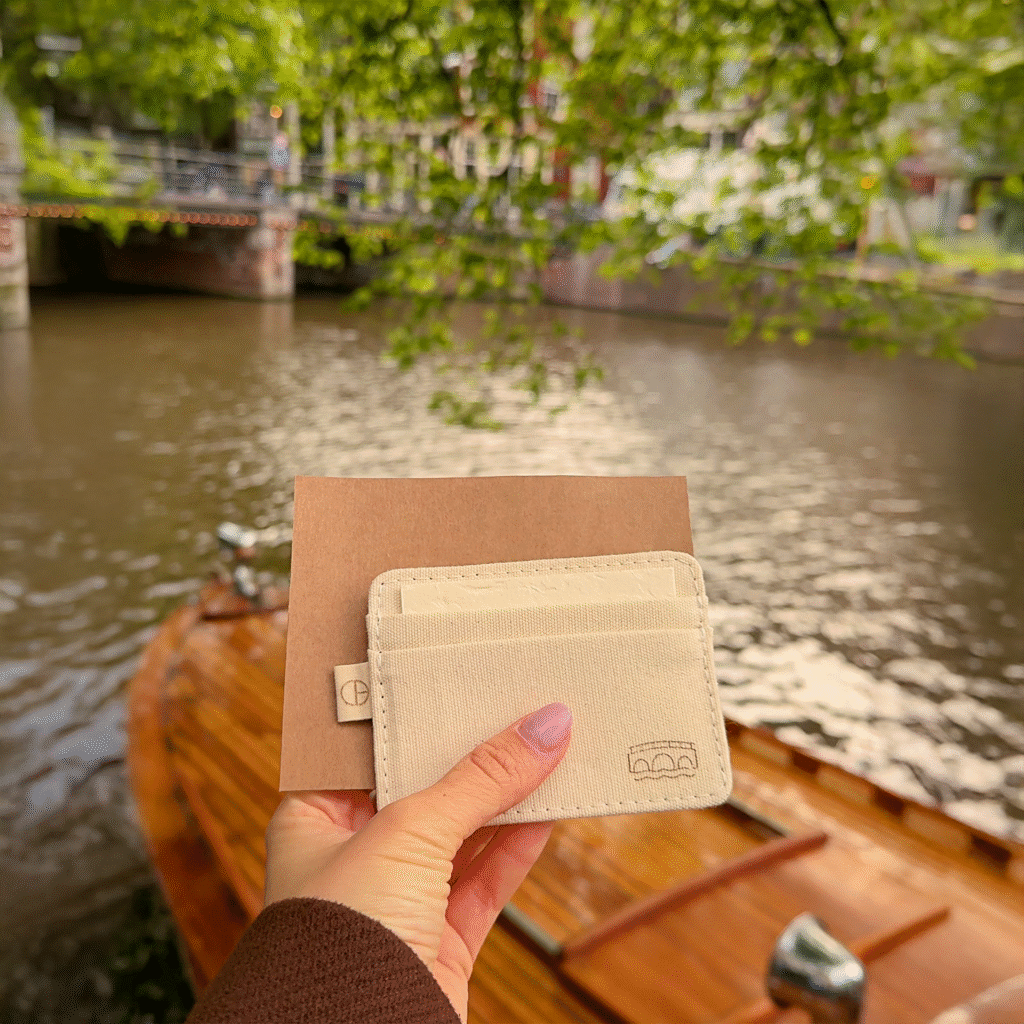Cream-colored CARDHΞ↓R cardholder held in front of an Amsterdam canal with a wooden boat.